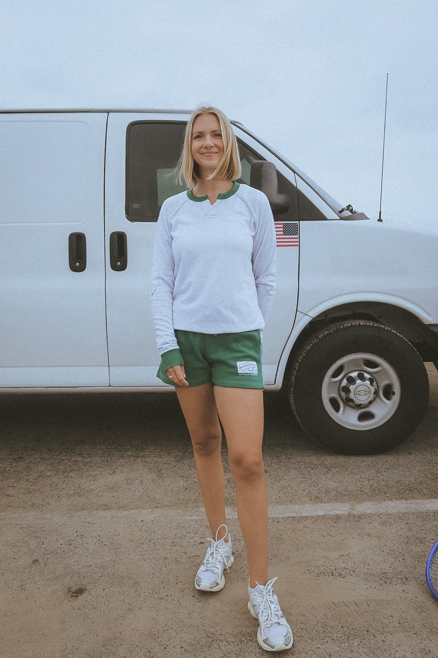 Person standing with a view of a building and another person in the distance by the ocean in california wearing ace the moon tennis henley