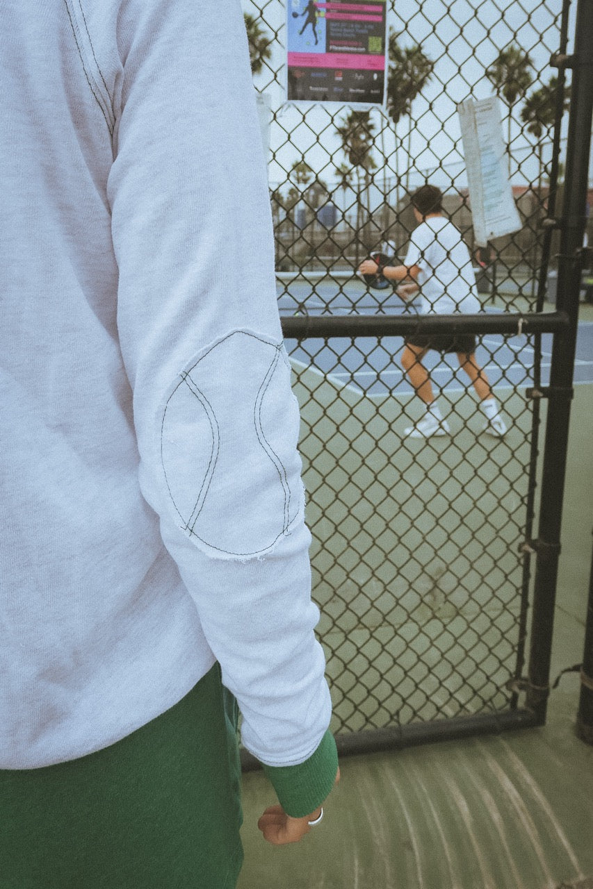 Person wearing a light gray hand knit tennis sweatshirt and green pants standing in front of a chain-link fence watching pickleball in california