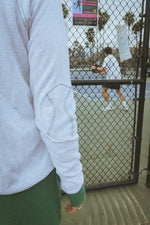 Person wearing a light gray hand knit tennis sweatshirt and green pants standing in front of a chain-link fence watching pickleball in california