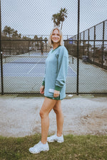 Woman in a light blue sweatshirt and white sneakers standing on a tennis court.