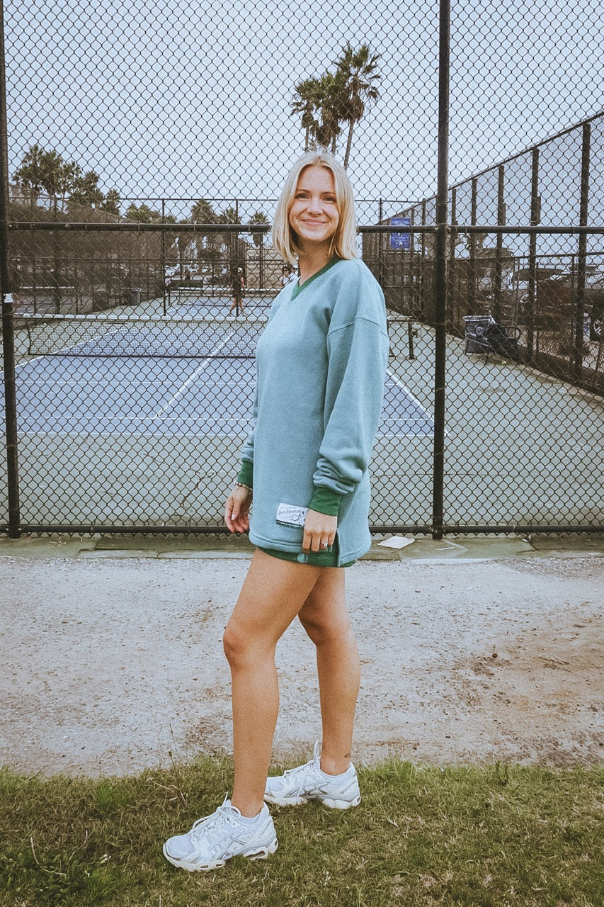 Woman in a light blue sweatshirt and white sneakers standing on a tennis court.