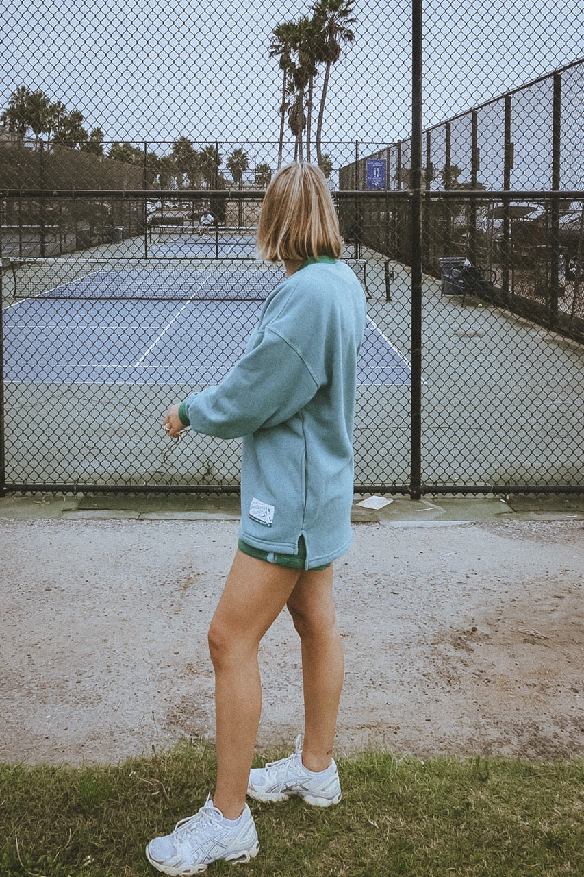 Person standing on a tennis court wearing a light blue sweatshirt and shorts.