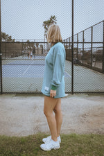 Person standing on a tennis court wearing a light blue sweatshirt and white sneakers.