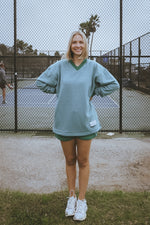 Person wearing a light green sweater and green shorts standing on a tennis court.