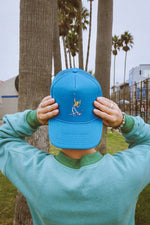Person holding a blue tennis trucker hat with a classic tennis player design in front of palm trees in california