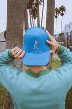 Person holding a blue tennis trucker hat with a classic tennis player design in front of palm trees in california