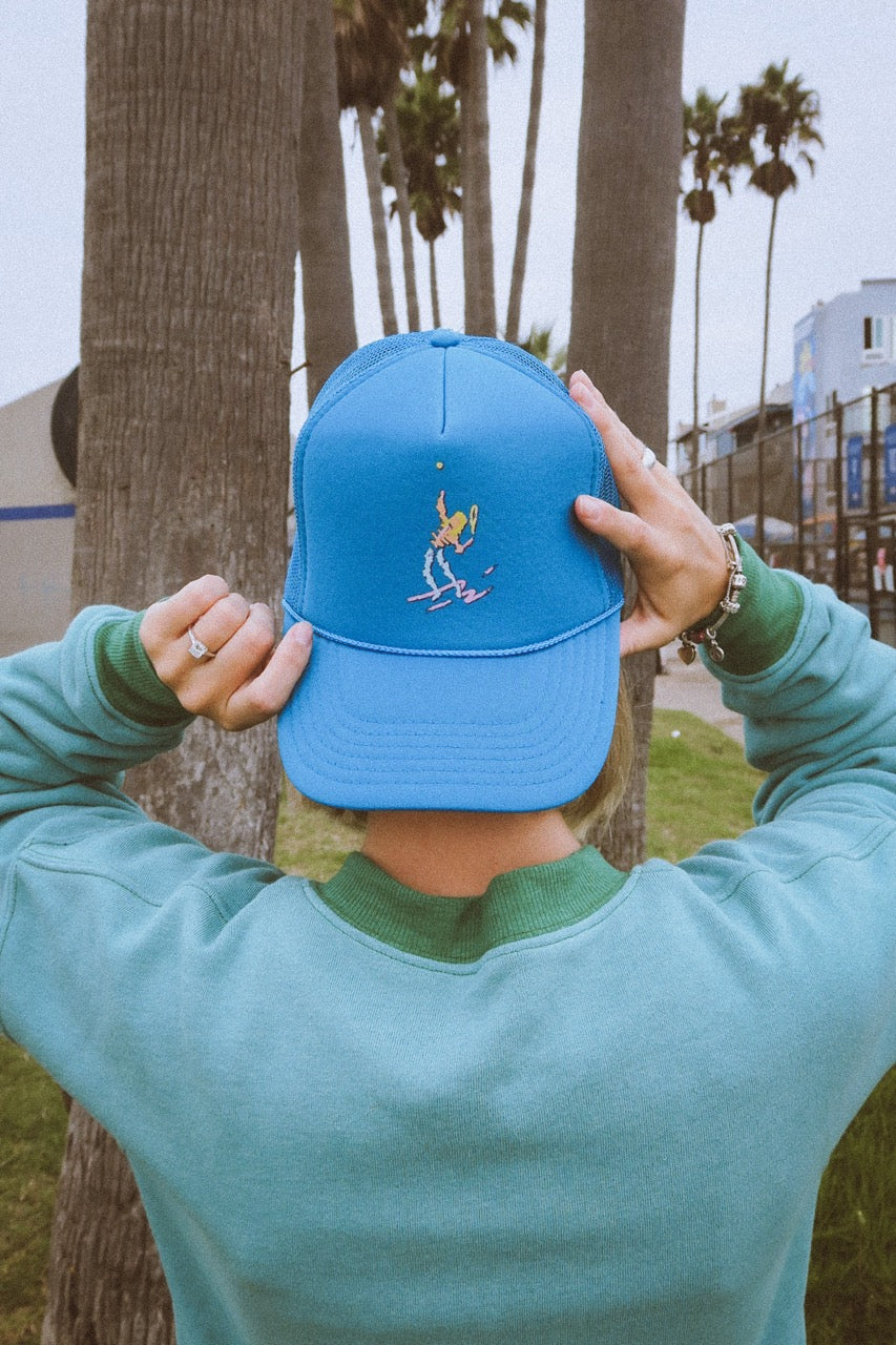 Person holding a blue tennis trucker hat with a classic tennis player design in front of palm trees in california