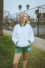 Person wearing a white sweater with green accents and green shorts standing on grass near a fence.