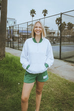Woman in a white sweatshirt and green shorts standing on grass with a tennis court in the background.