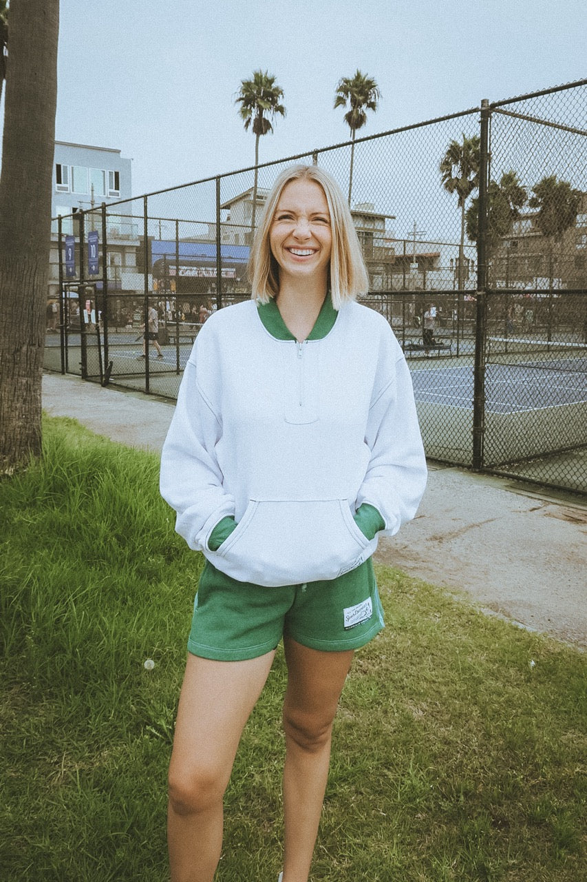 Woman in a white sweatshirt and green shorts standing on grass with a tennis court in the background.