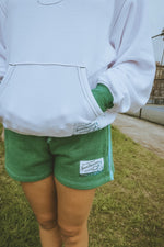 Person wearing a white hoodie and green shorts with brand labels, standing on grass.