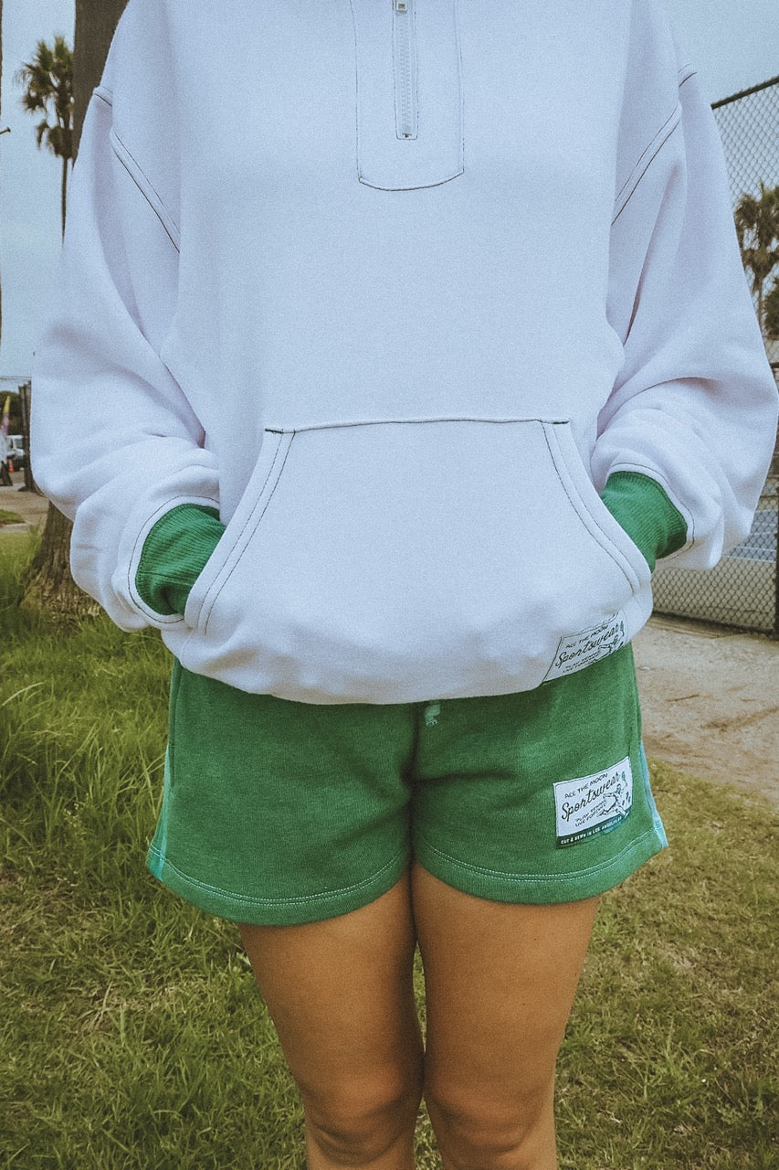 Person wearing a white tennis zip sweatshirt and green shorts with a ace the moon brand logo outdoors.