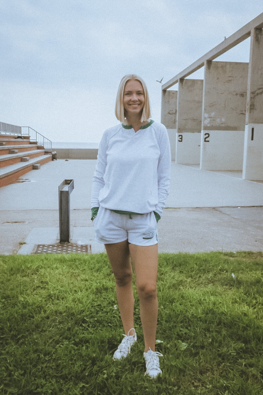 Person standing with a view of a building and another person in the distance by the ocean in california wearing ace the moon tennis henley