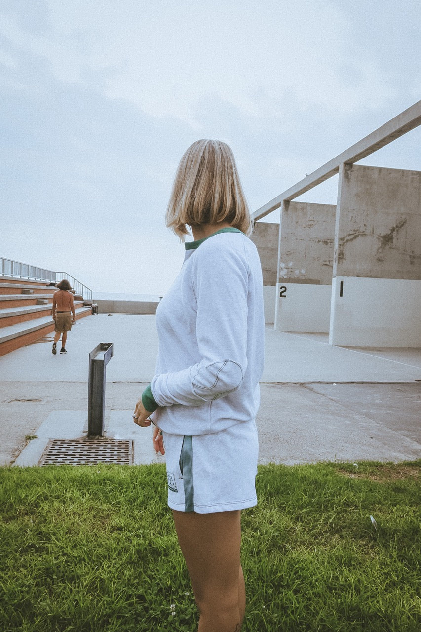 Person standing with a view of a building and another person in the distance by the ocean in california wearing ace the moon tennis henley