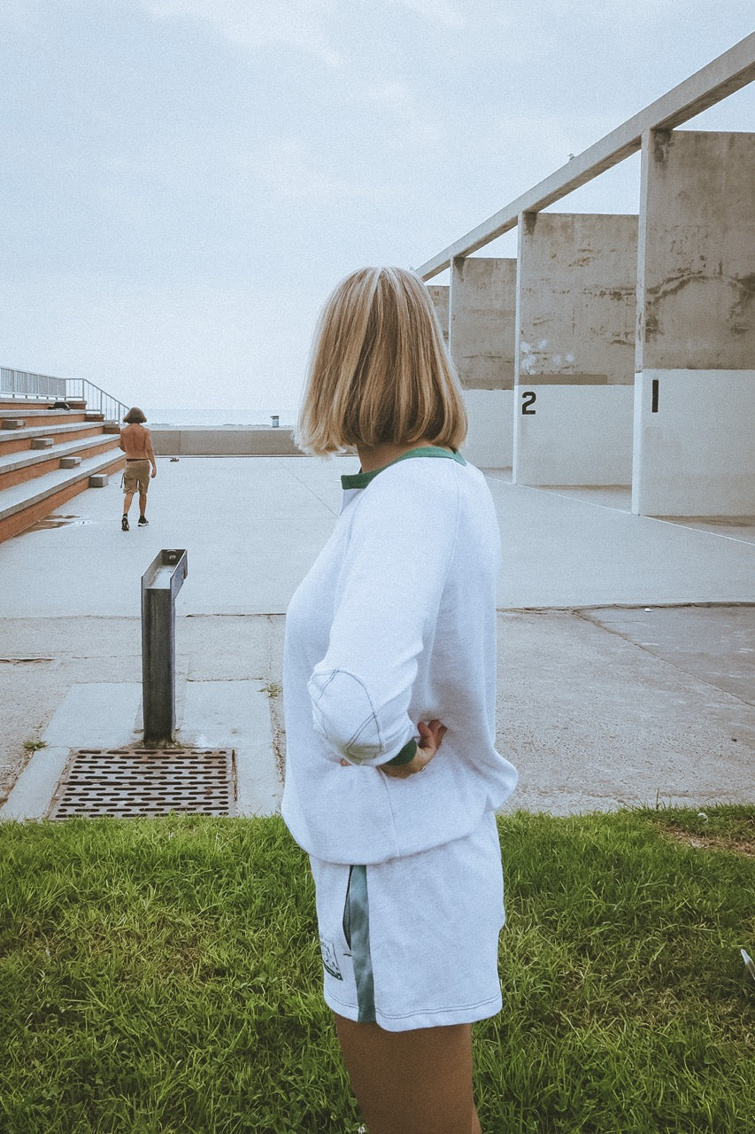 Person standing with a view of a building and another person in the distance by the ocean in california wearing ace the moon tennis henley