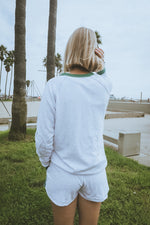 Person wearing a white sweatshirt and shorts standing by palm trees near a body of water.