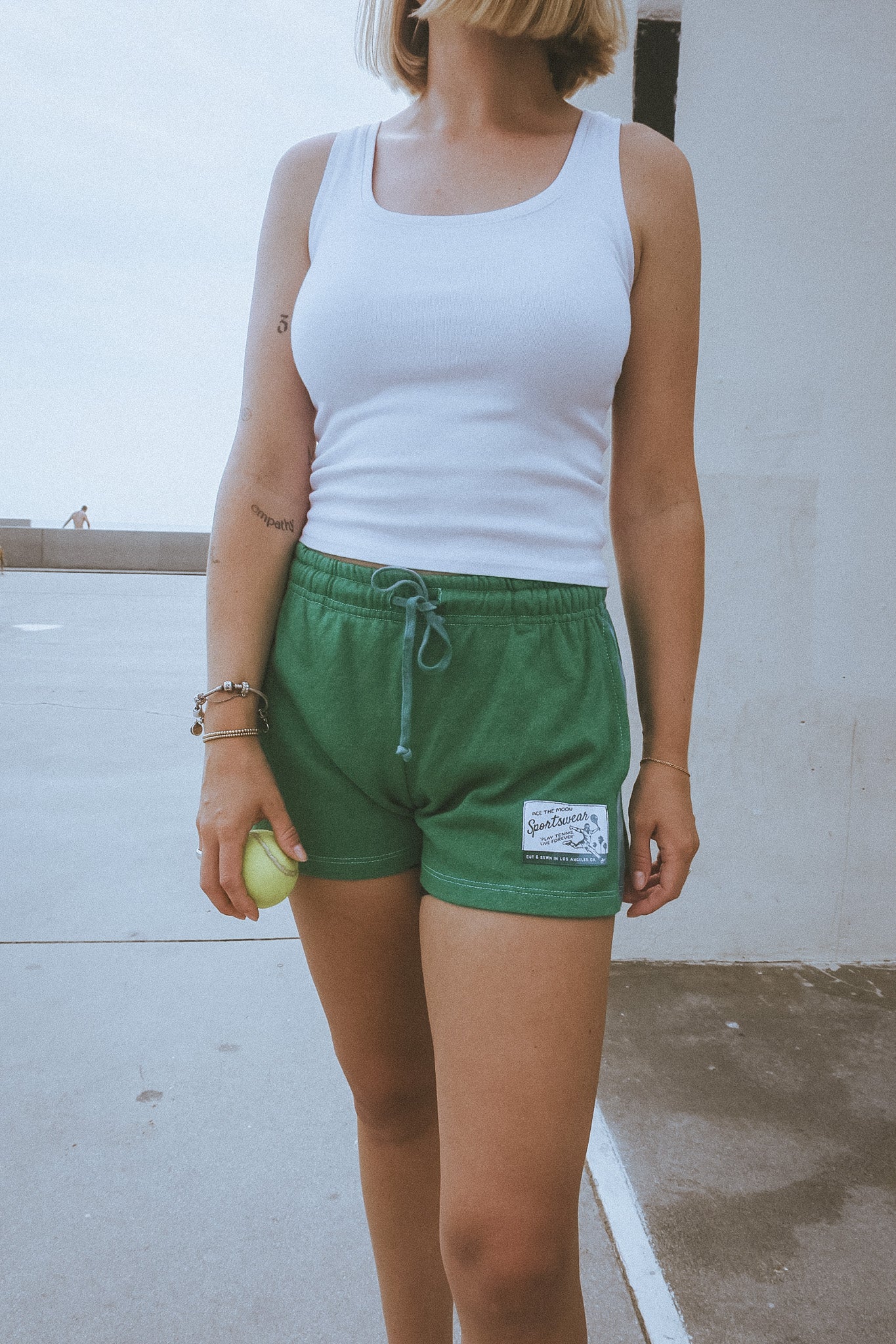 Person wearing a white tank top and green shorts with a logo, standing against a light-colored wall.