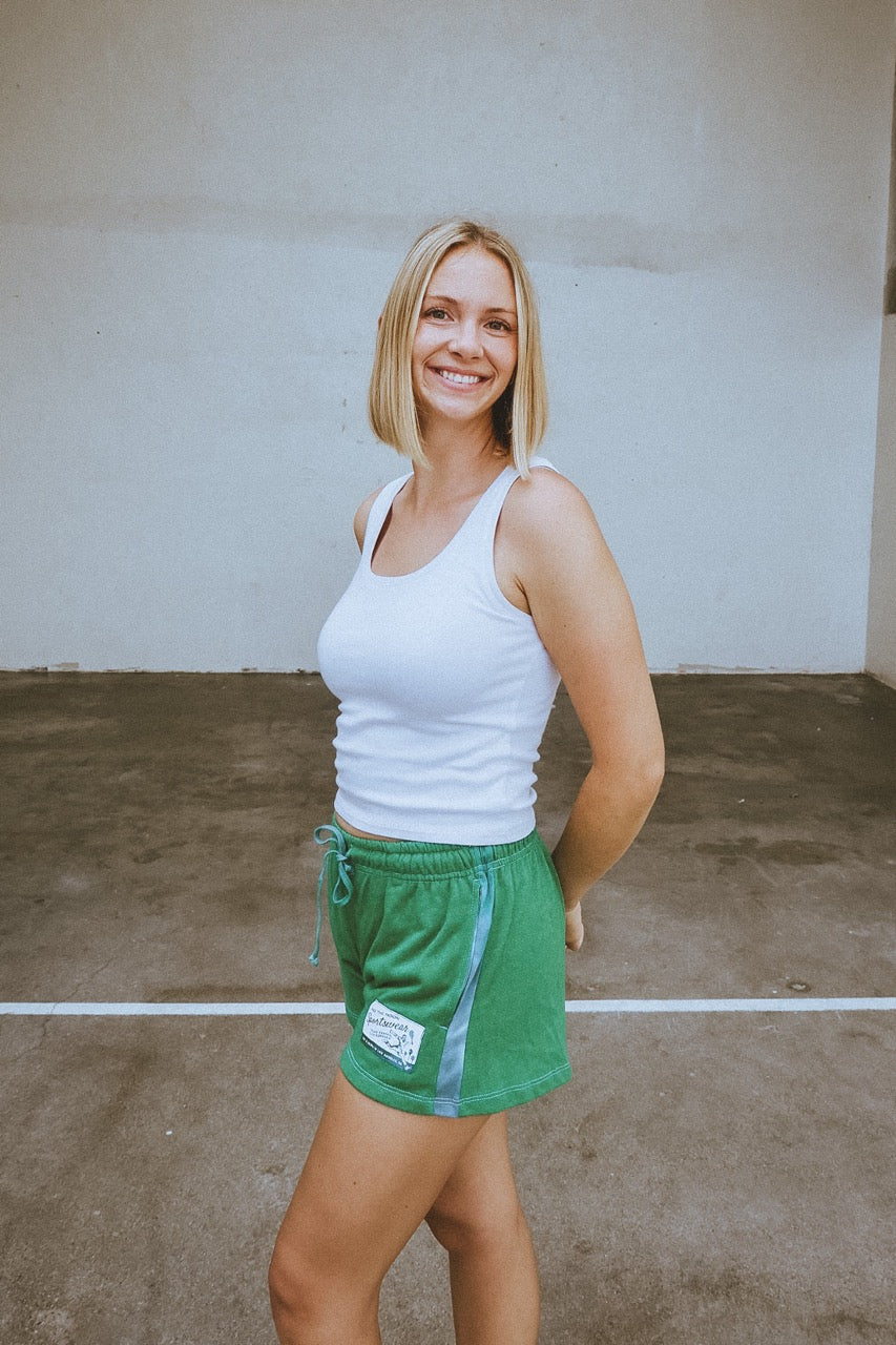Woman wearing a white tank top and green shorts standing against a plain wall.