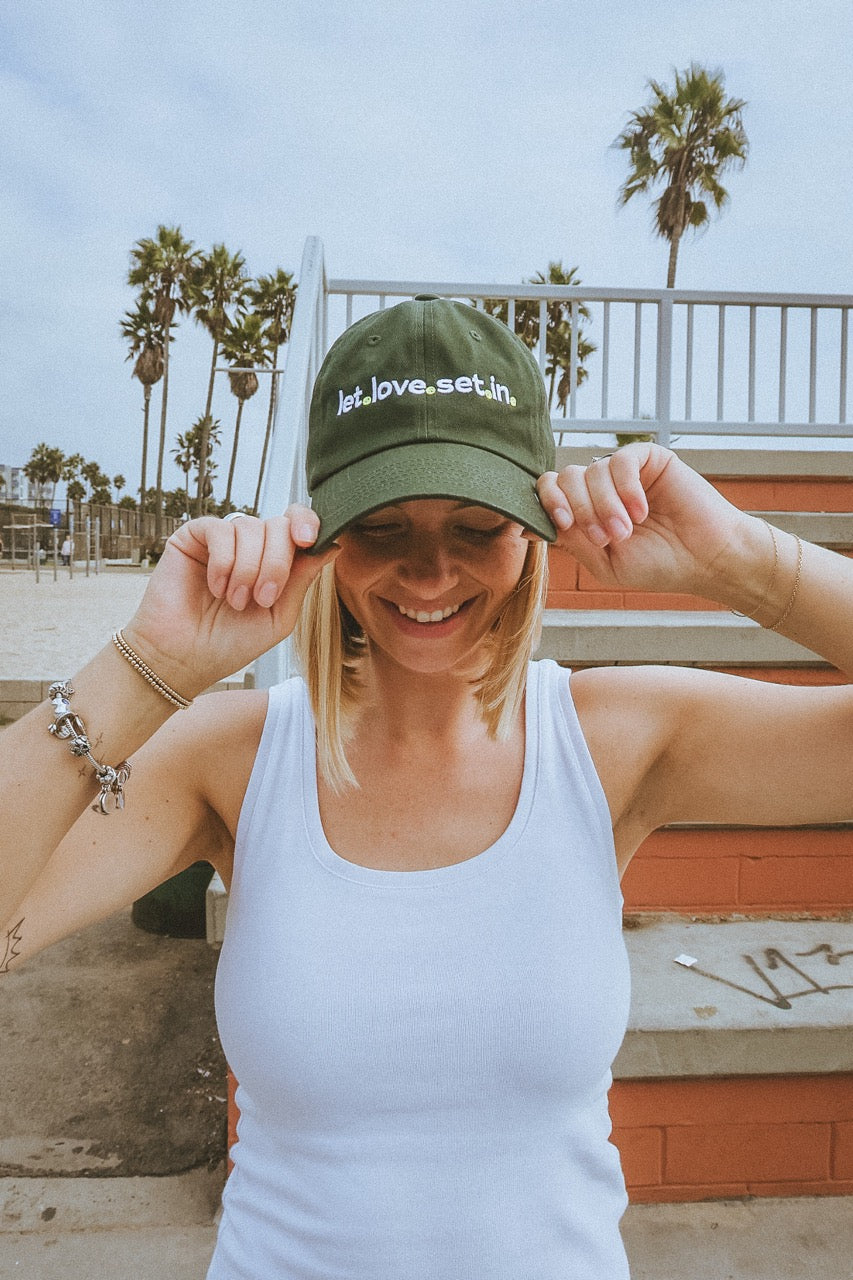 Woman wearing a green tennis dad hat by ace the moon with text with text that says let love set in, standing outdoors with palm trees in the background