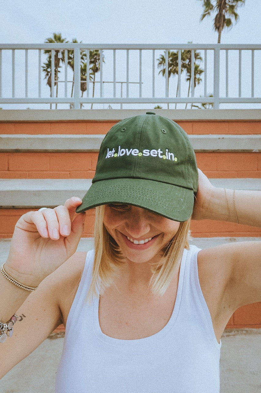 Woman wearing a green tennis dad hat by ace the moon with text with text that says let love set in, standing outdoors with palm trees in the background