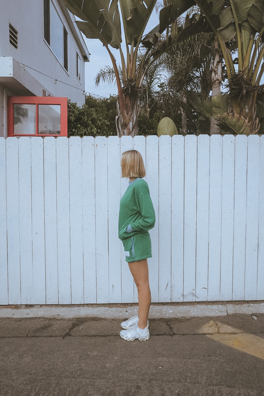 Person in a green outfit standing in front of a white fence with palm trees in the background