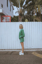 Person in a green outfit standing in front of a white fence with palm trees in the background