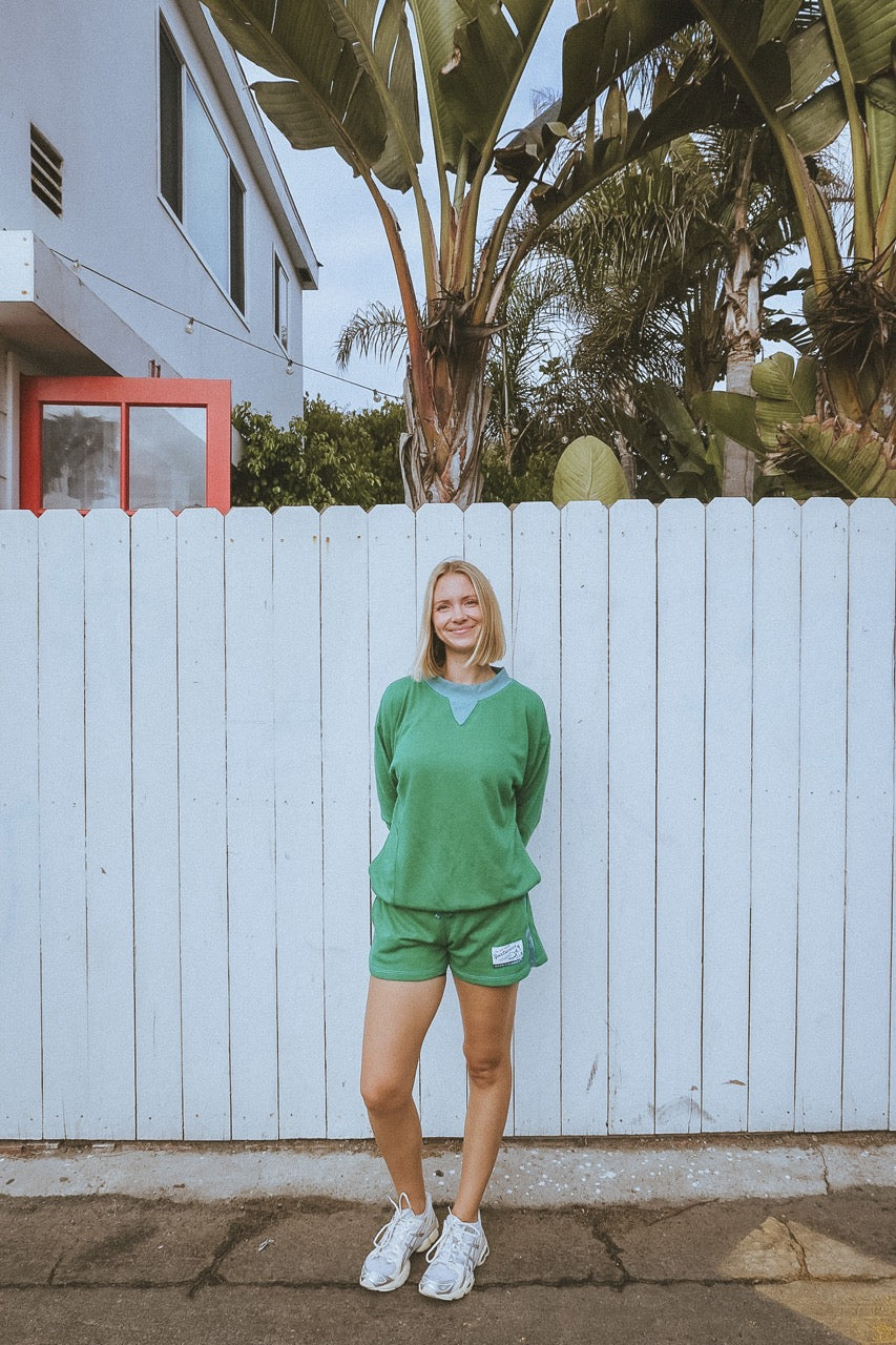 Person wearing a green outfit standing in front of a white fence with palm trees in the background.