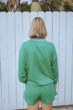 Person wearing a green outfit standing in front of a white fence with trees in the background