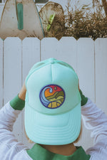 Person wearing a light blue tennis trucker hat with a colorful logo against a white picket fence.