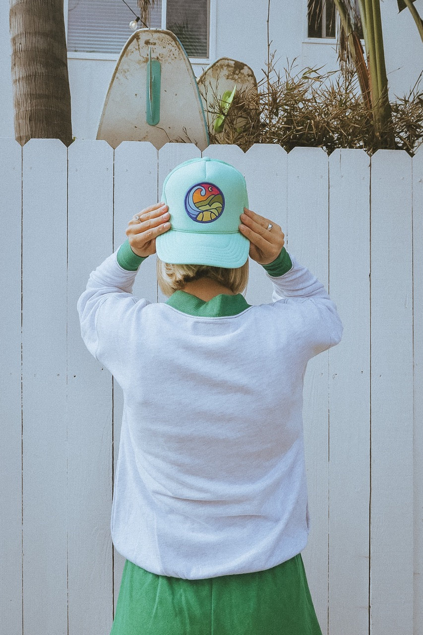 Person wearing a light blue tennis trucker hat with a colorful logo against a white fence background
