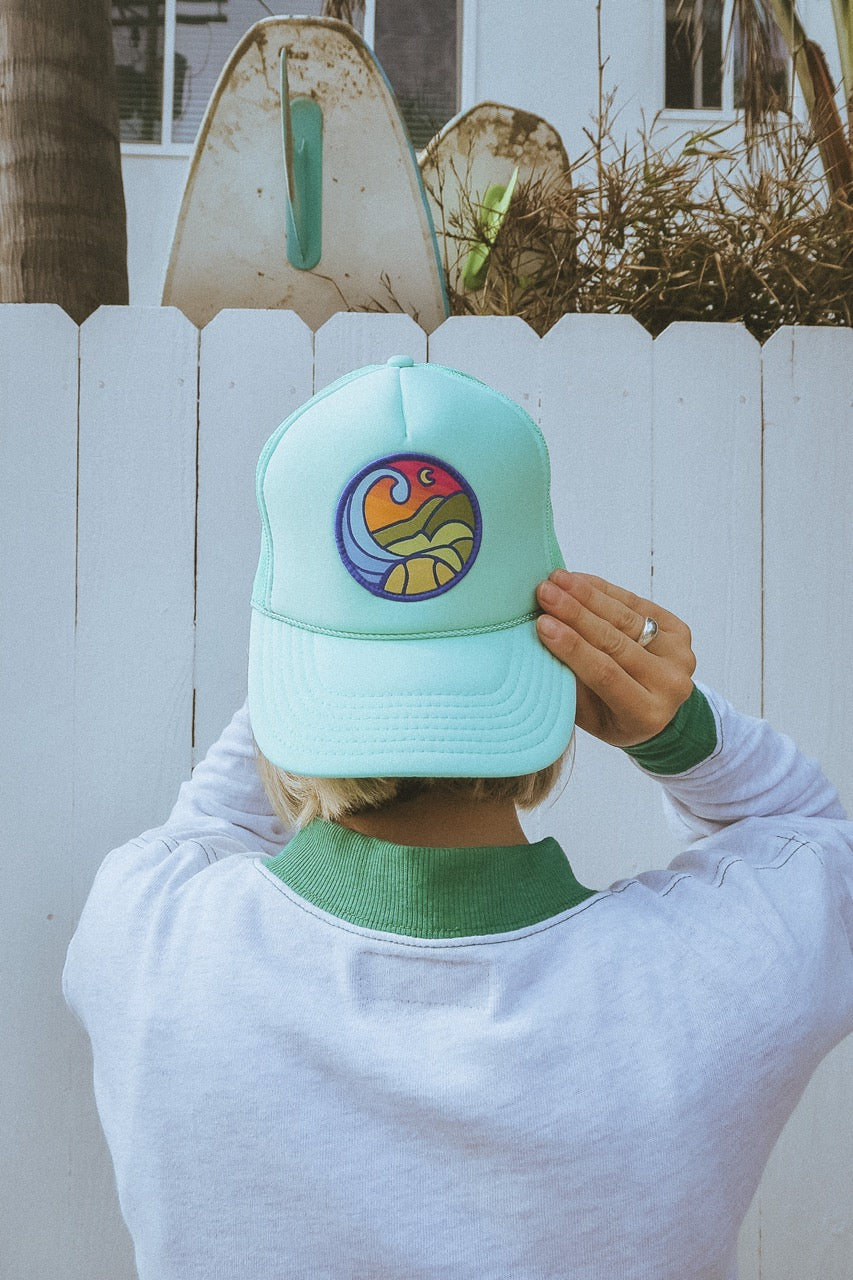 Person wearing a light blue tennis trucker hat with a colorful logo against a white fence background