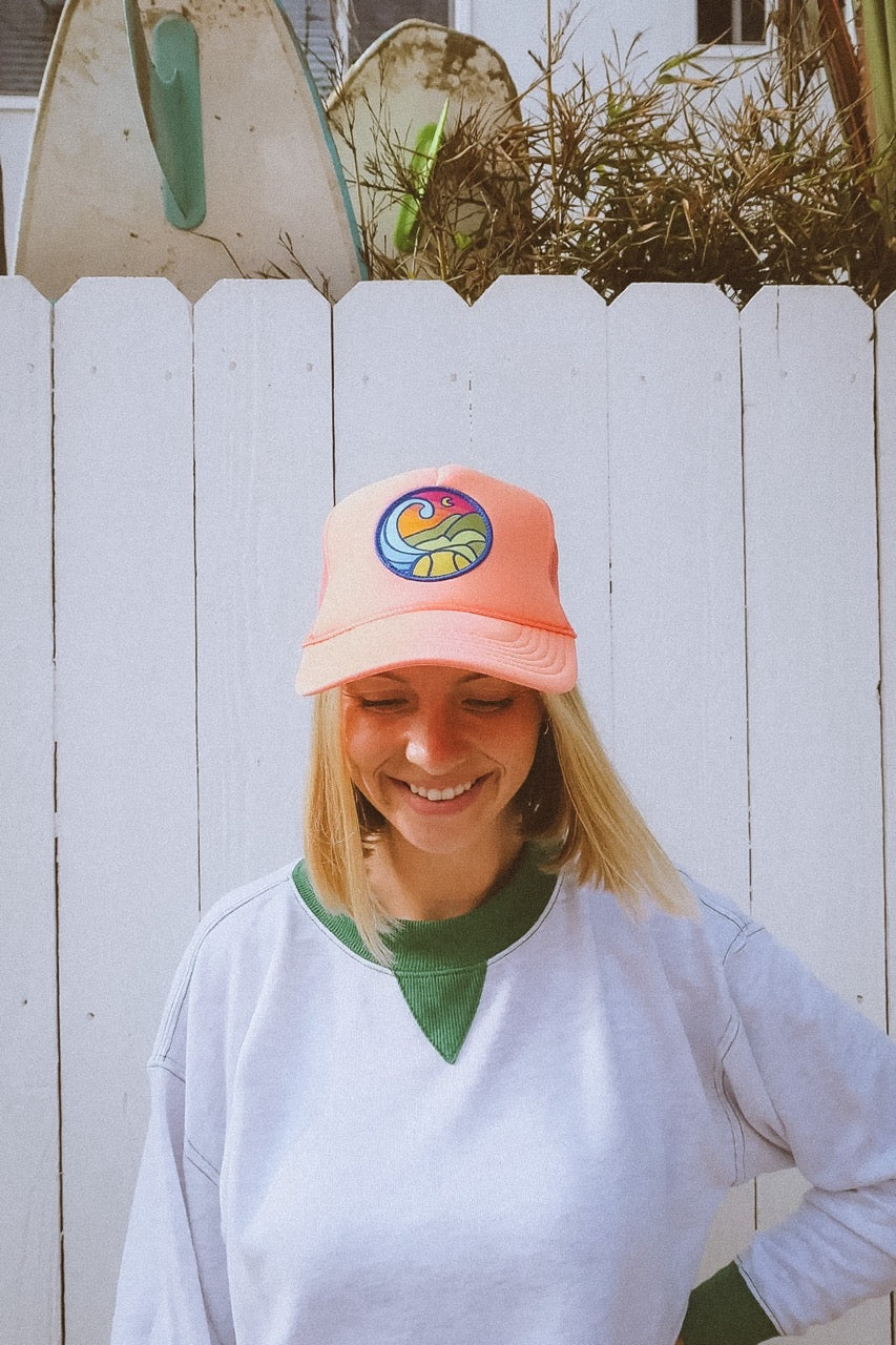 Person wearing a coral colored tennis trucker hat with a colorful logo against a white wooden fence.