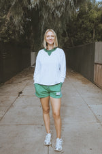 Woman in a white sweatshirt and green shorts standing on a sidewalk with trees in the background