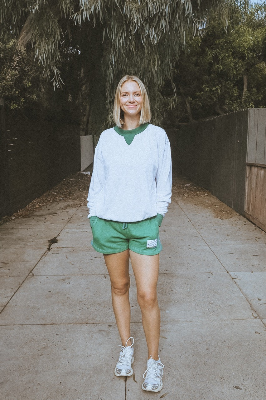 Woman in a white sweatshirt and green shorts standing on a sidewalk with trees in the background
