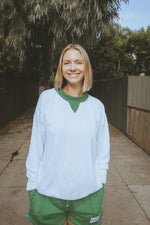 Person wearing a white sweater and green pants standing outdoors with trees in the background