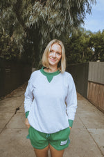 Woman wearing a white tennis sweatshirt and green shorts standing outdoors with trees in the background