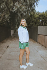 Woman in a white sweatshirt and green shorts standing on a sidewalk with trees in the background