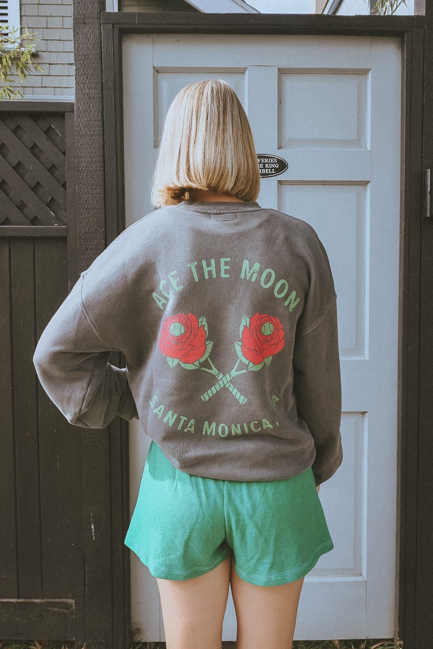 girl wearing a vintage black washed tennis crewneck sweatshirt by ace the moon tennis apparel brand that has text and rose shaped tennis rackets and santa monica, ca