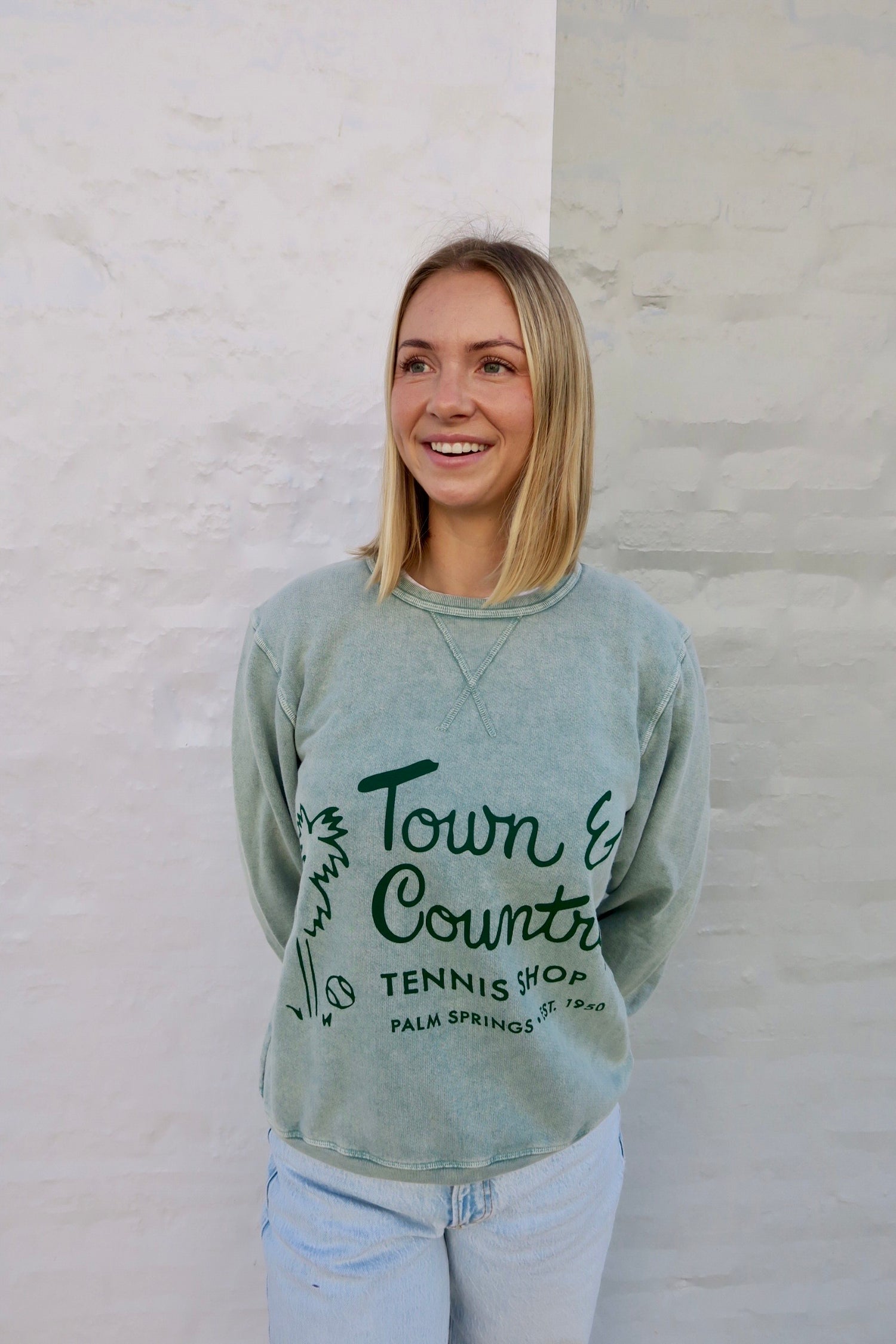 Woman wearing a green sweatshirt with text standing against a light-colored wall.