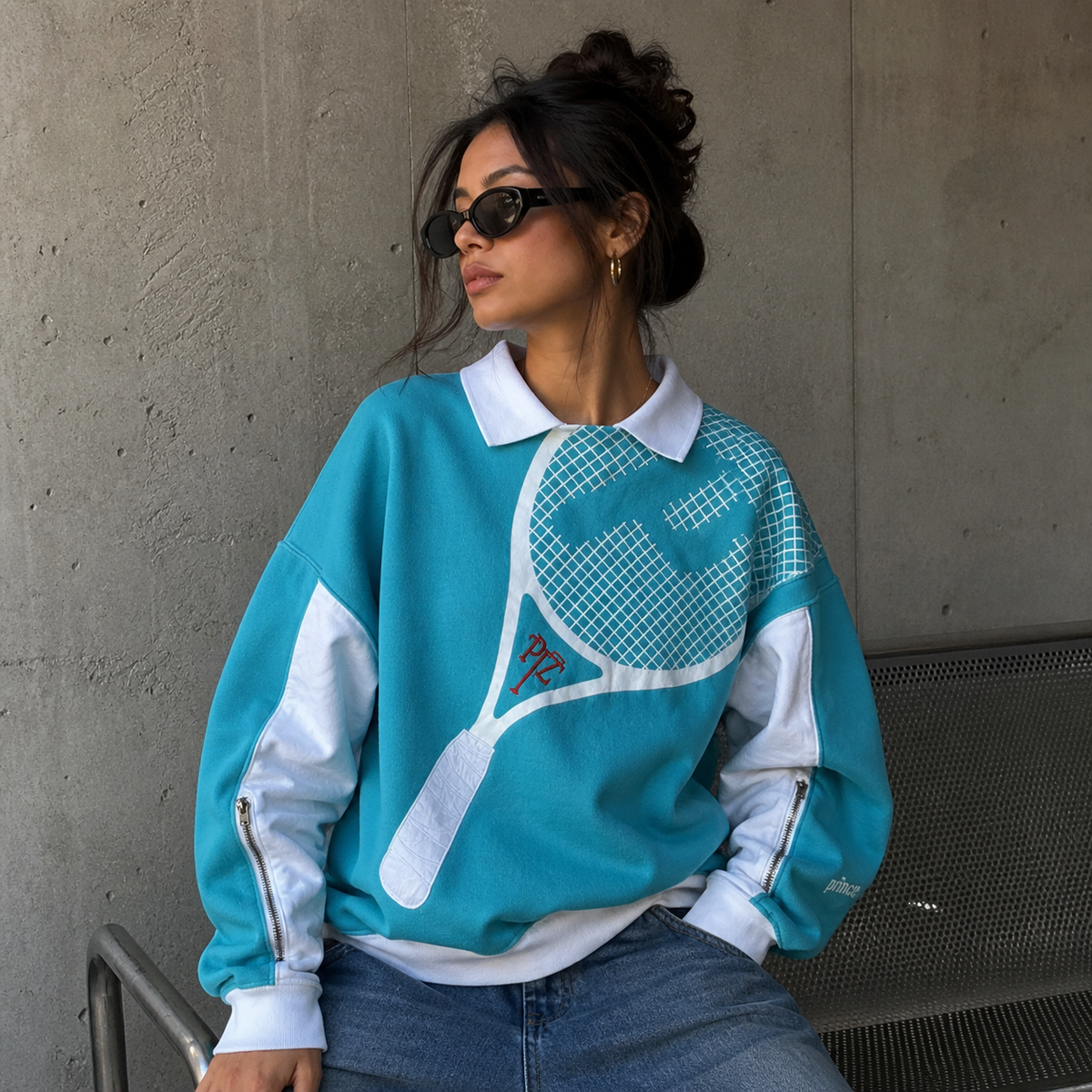 Woman wearing a teal and white sweatshirt with a tennis racket design against a gray wall.