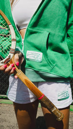 Person wearing a green hoodie with a logo, holding a wooden tennis racket.