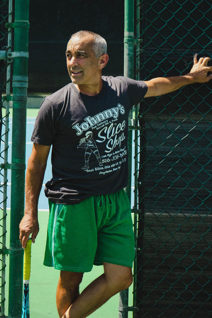 Man wearing a black t-shirt with text and green shorts leaning against a fence.