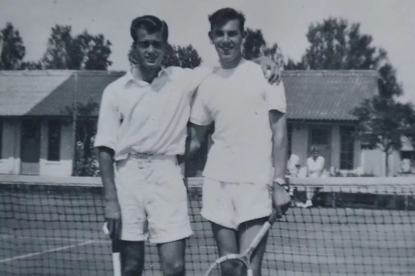 Two men standing on a tennis court holding rackets, with a building and trees in the background.