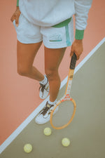 Person on a tennis court with a racket and balls, wearing a white outfit with green accents.