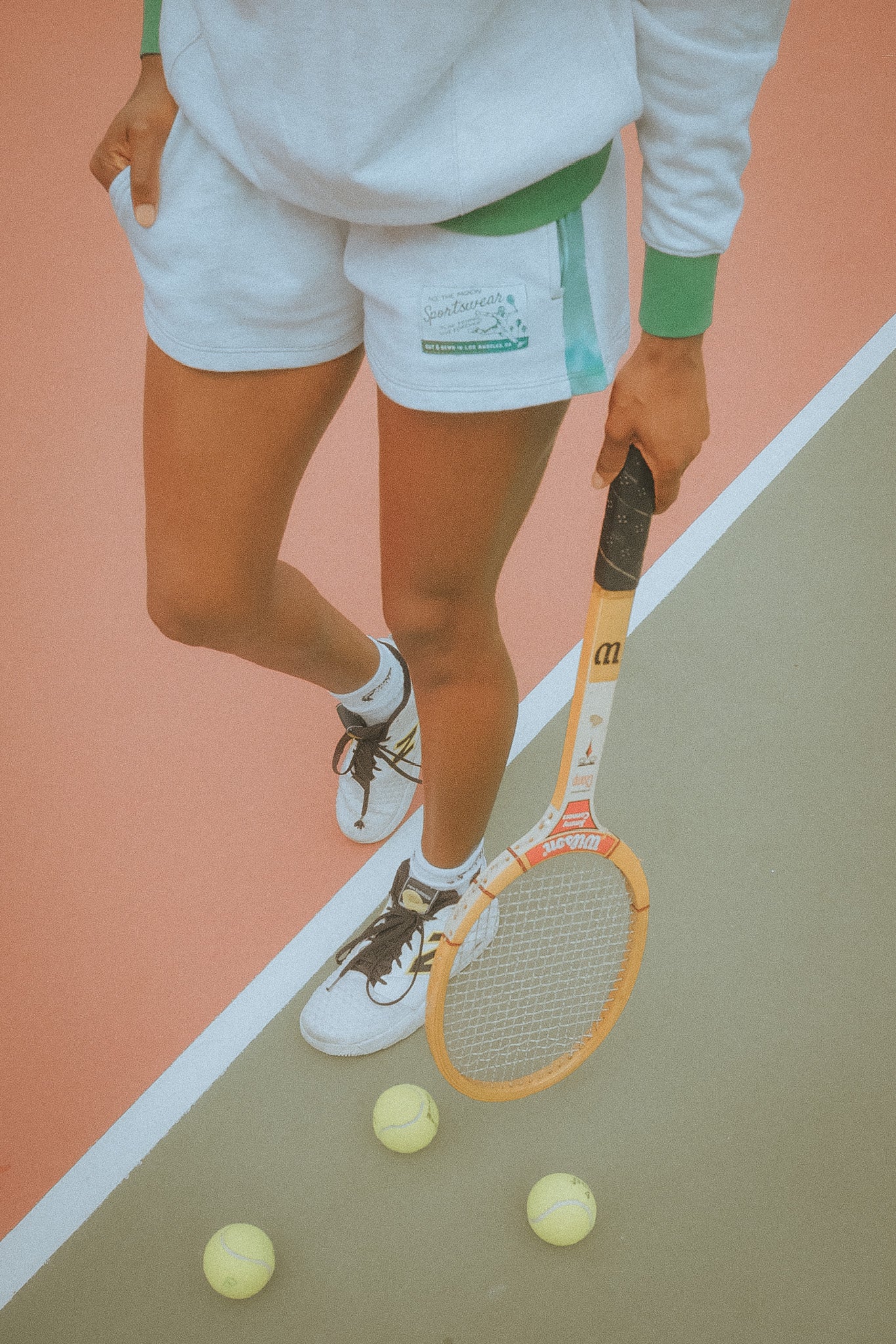Person on a tennis court with a racket and balls, wearing a white outfit with green accents.