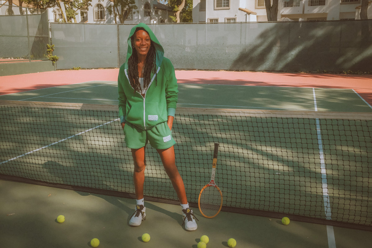 Person in green outfit standing on a tennis court with tennis balls and racket.