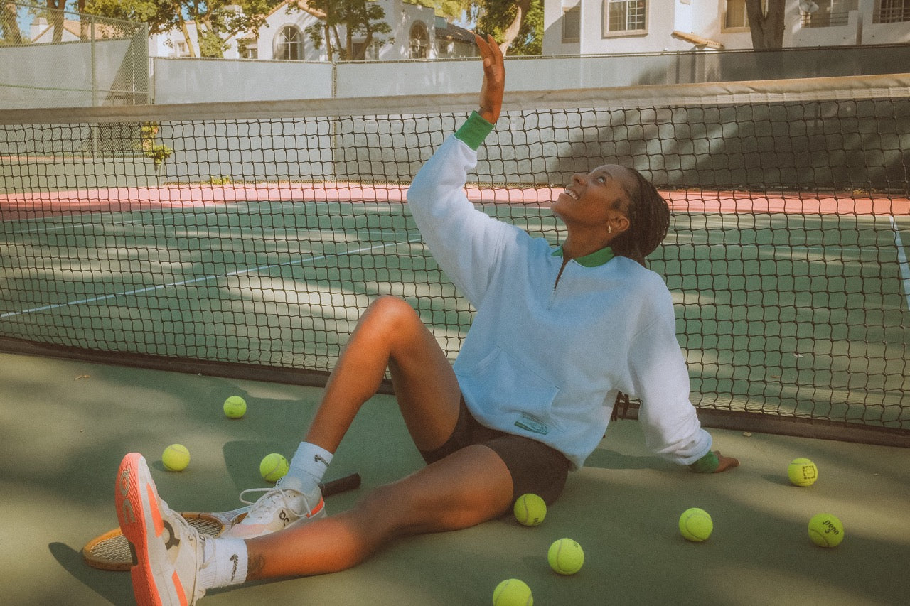 Person sitting on a tennis court surrounded by tennis balls, with a net in the background.