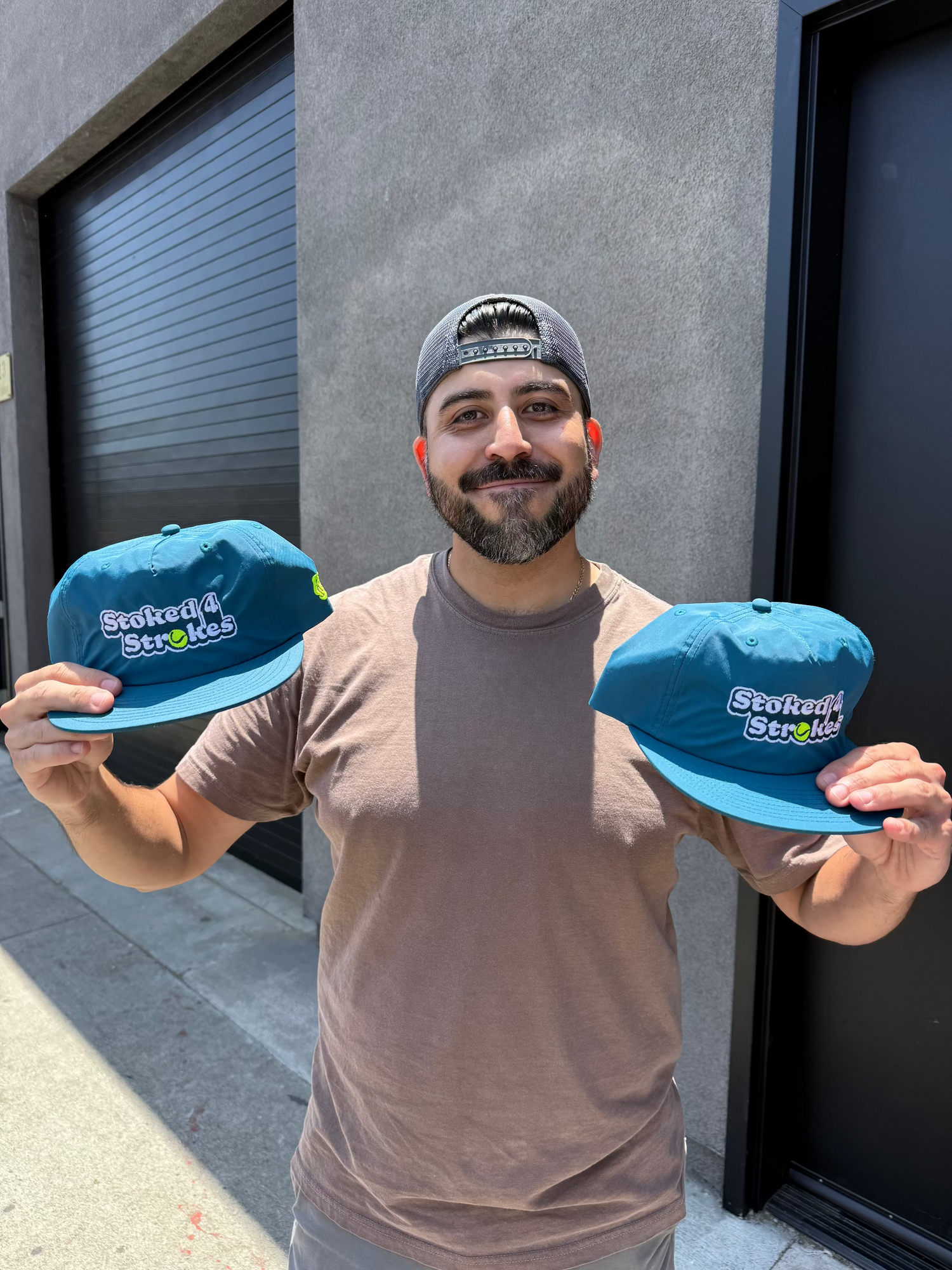 Man holding two blue hats with visible branding against a gray wall.