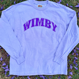 "WIMBY" long sleeve tee in vintage purple/lavender color with collegiate purple font laying on the ground surrounded by purple flower petals. made by Ace the moon premium tennis apparel brand
