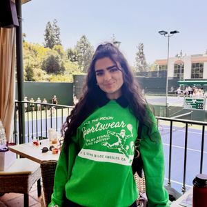 ace the moon tennis sportswear premium hand-knit sweater in green worn by a female tennis player standing in front of an outdoor tennis court